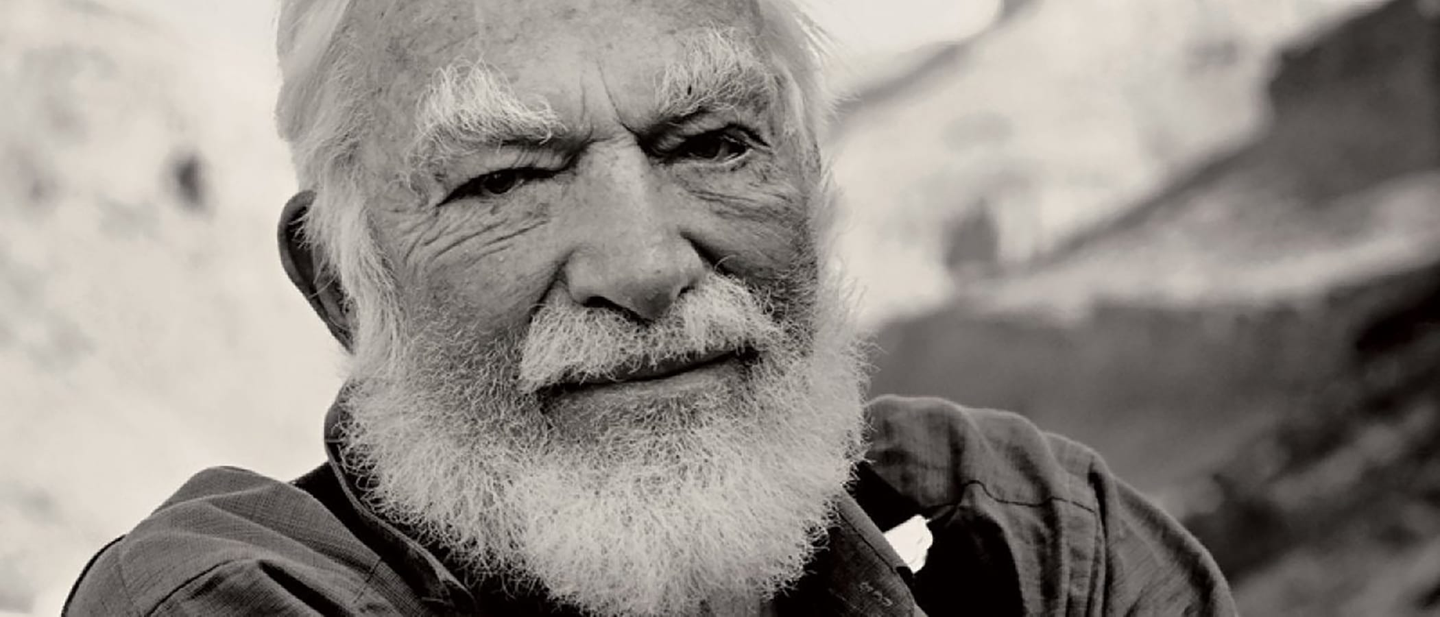 A bearded white haired person from the shoulders up with mountains out of focus in the background.