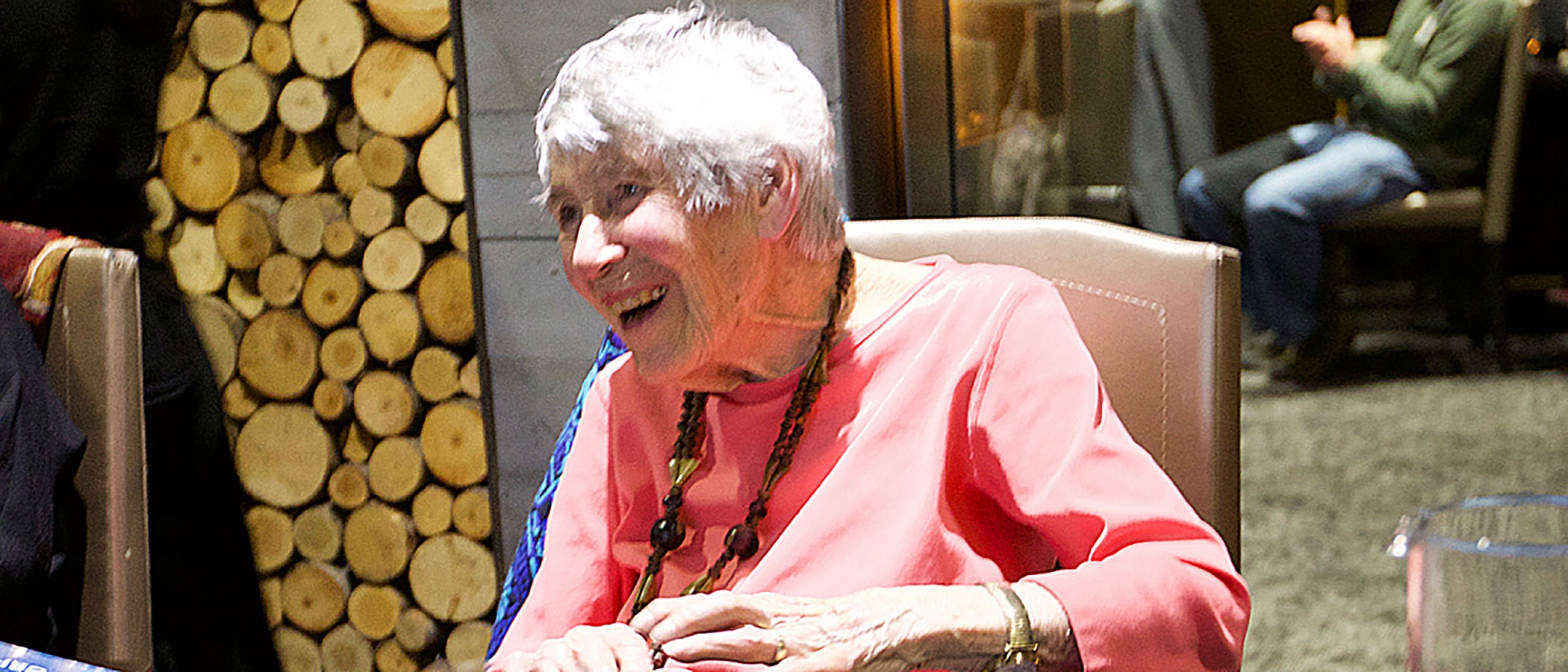 A white haired person in a pink shirt sits in a chair, behind them an orderly stack of firewood rounds can be seen.