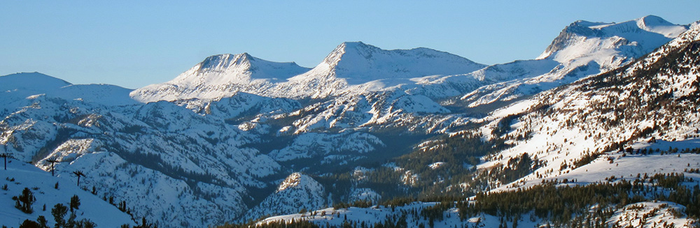 A view across an open snow-covered mountain valley spotted with pine trees to three alpine peaks above the tree line with rocky crags and the bright light and blue shadows of a sunset under a bright blue sky, with the middle peak being Mount Andrea Lawrence.