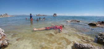 A boy wearing goggles and smiling lays in Mono Lake near the shore, floating on his back. Clear blue skies and clear greenish blue water stretch out above, and more people are swimming.