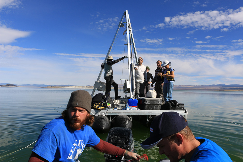 Seven researchers out on Mono Lake on a pontoon platform with tall metal hoisting structure and lots of research equipment on it, being pulled by a smaller motor boat.