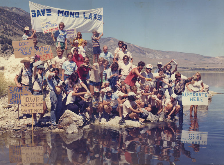 Group of about 50 people with hand-painted signs and Mono Lake T-shirts and many with bike helmets on pouring water from bottles and vials into Mono Lake.