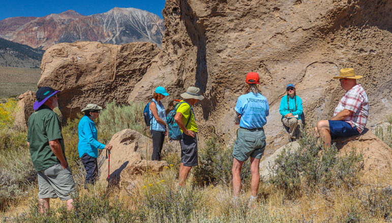 Seven people wearing hiking gear gather around a guide leaning on an unusual rock in a rocky and sunny outdoor setting.