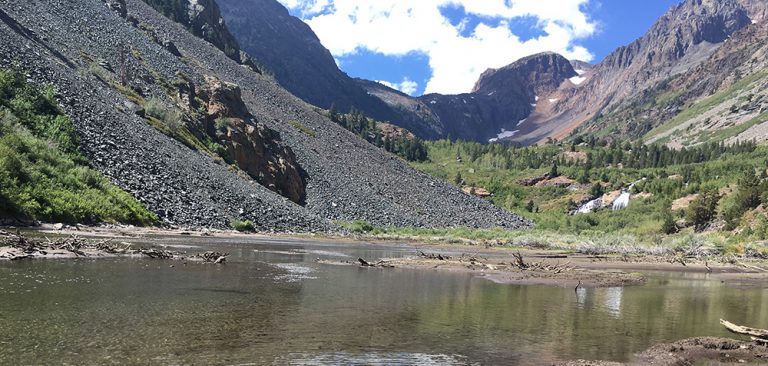 Water flows in front of a scree slope, further up a u shaped valley trees cover the bottom and towering rock faces line the edge, bright clouds cover part of the blue sky.