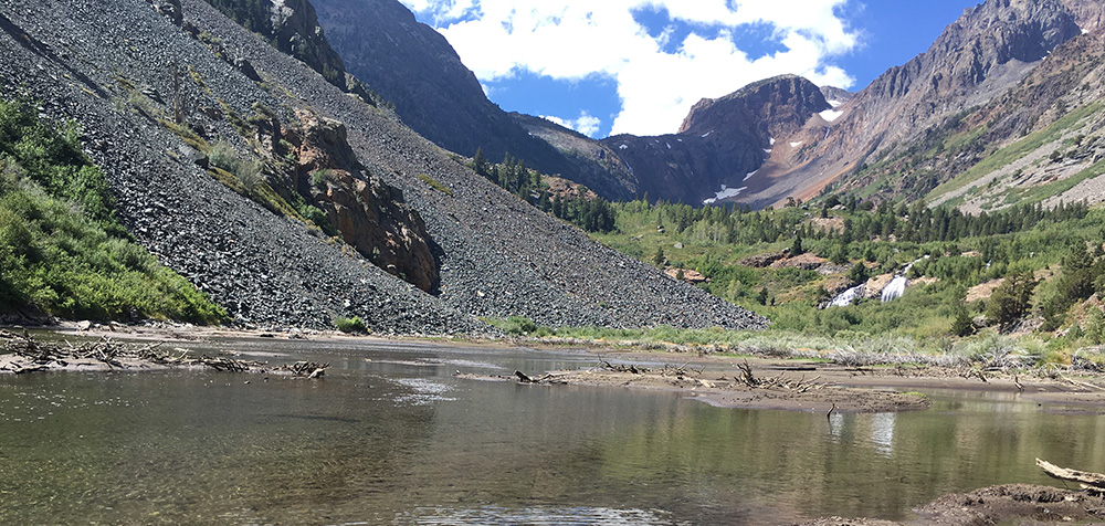 Water flows in front of a scree slope, further up a u shaped valley trees cover the bottom and towering rock faces line the edge, bright clouds cover part of the blue sky.