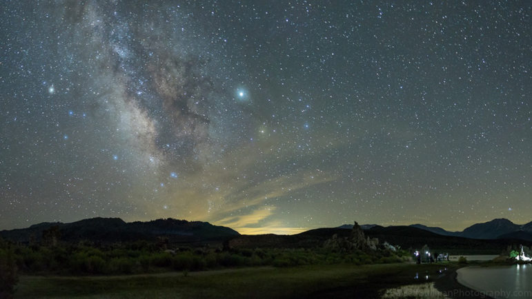 The milky way visible behind mountain outlines, in the foreground photographers are moving around with lights on the shore of a lake with tufa formations.