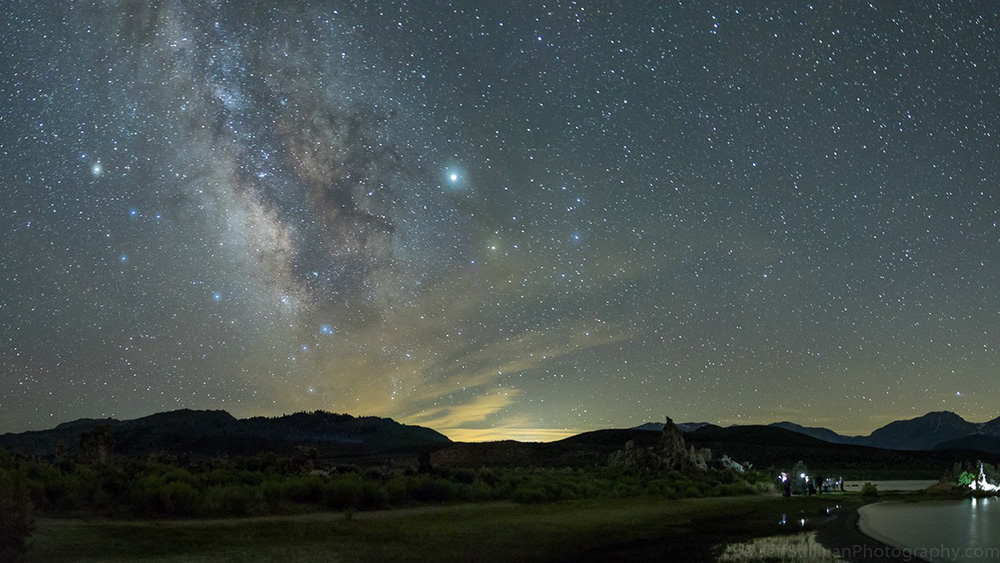 The milky way fills the sky while lights of people on the shore among tufa are seen below the horizon.