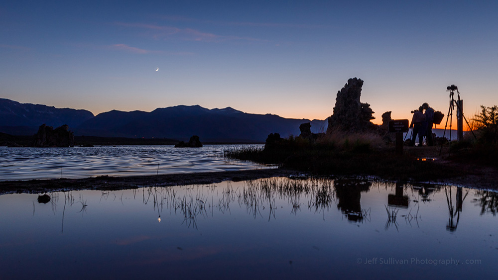 Two photographers stand in the growing dark next to their tripods among tufa on the shore of Mono Lake silhouetted against the fading orange of sunset, a sliver of a moon hangs above the mountains in the distance.