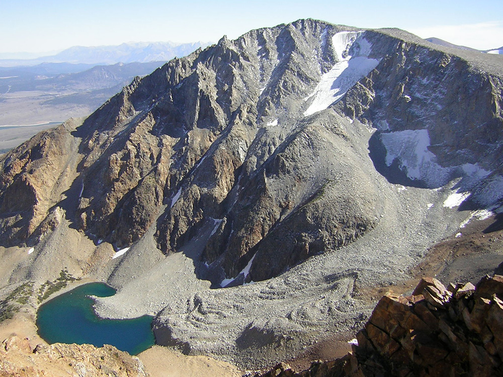 A small kidney shaped blue lake sits at the bottom of a rough scree covered peak with glaciers in the shade near the top.