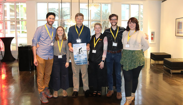 Six staff members of the Mono Lake Committee stand together indoors, smiling. Geoff McQuilkin holds a poster for the Wild and Scenic Film Festival.