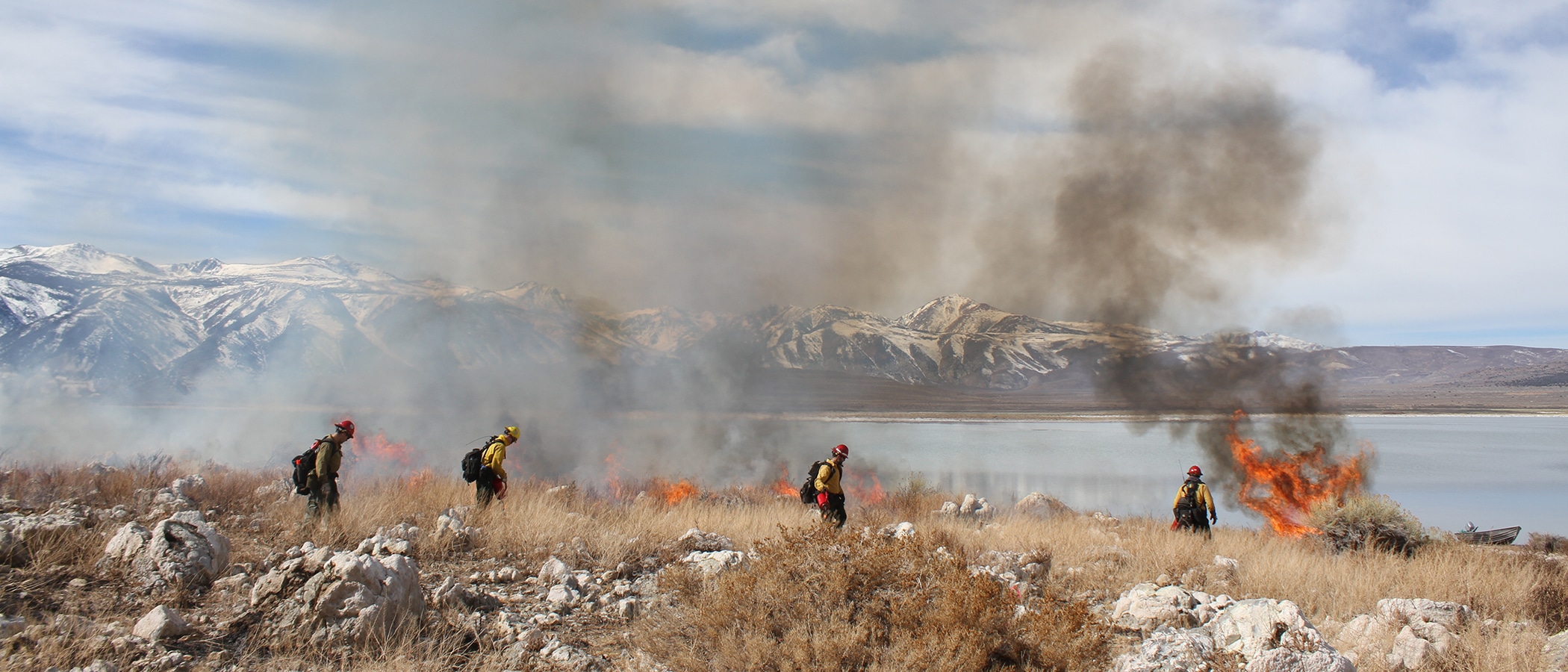 Wildland firefighters walking across rocky dry brush with flames and smoke rising against the backdrop of Mono Lake and the Sierra Nevada.