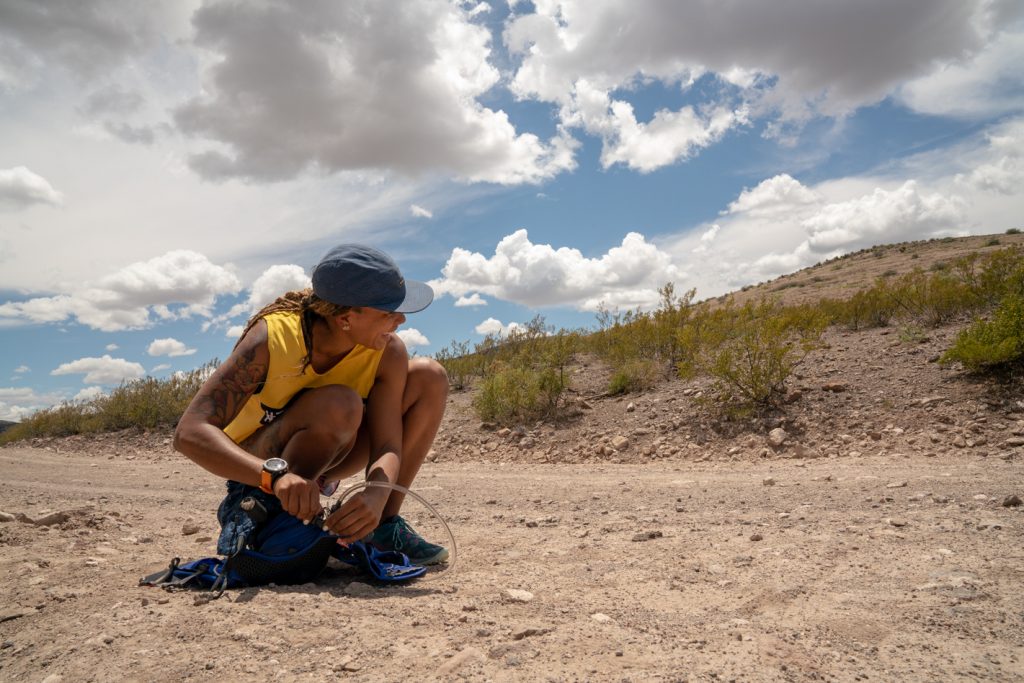 A runner wearing a yellow shirt crouches while taking a break along a dirt road in the desert with blue skies and white clouds in the background.