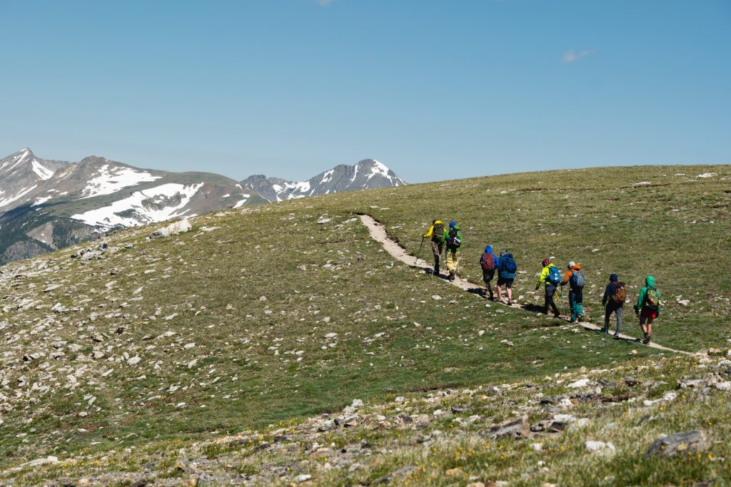 Eight hikers walk along a trail in the mountains wearing brightly colored outdoor gear.