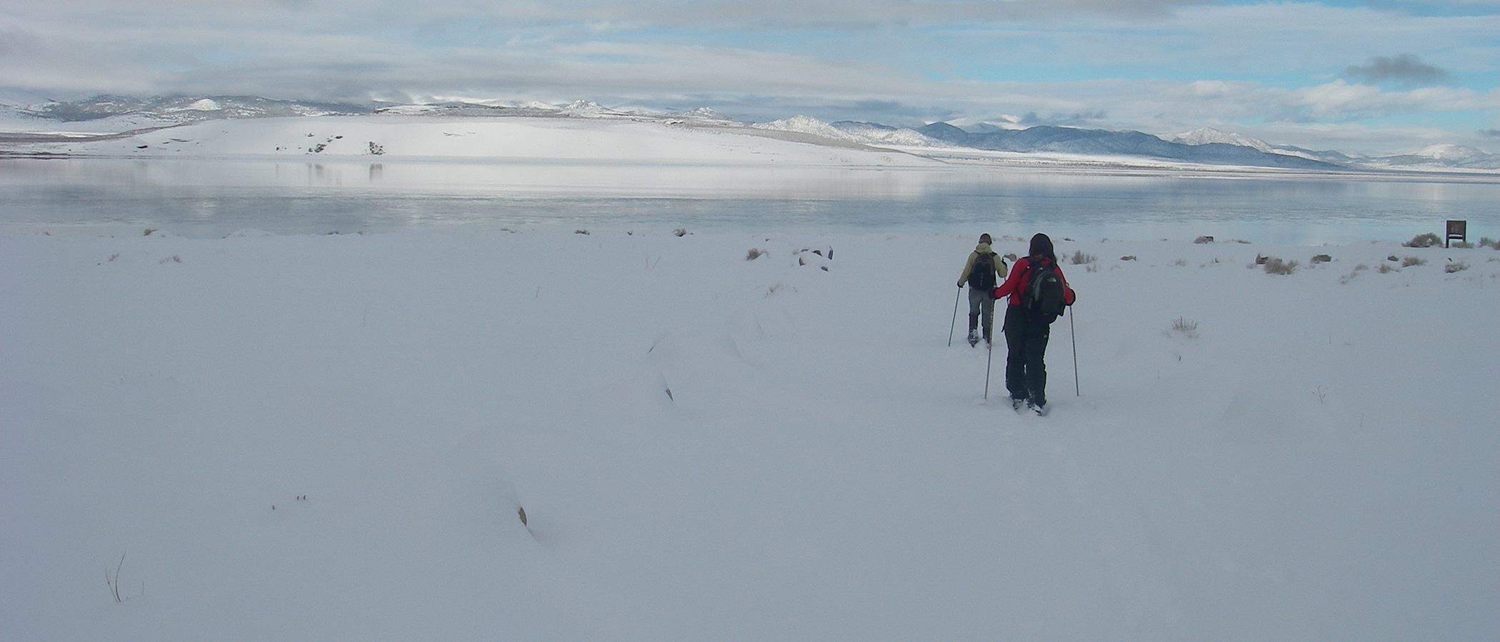 Two figures with ski poles are in motion in a snow field. Ahead of them, Mono lake is highly reflective of the white mountains and blue gray clouds.