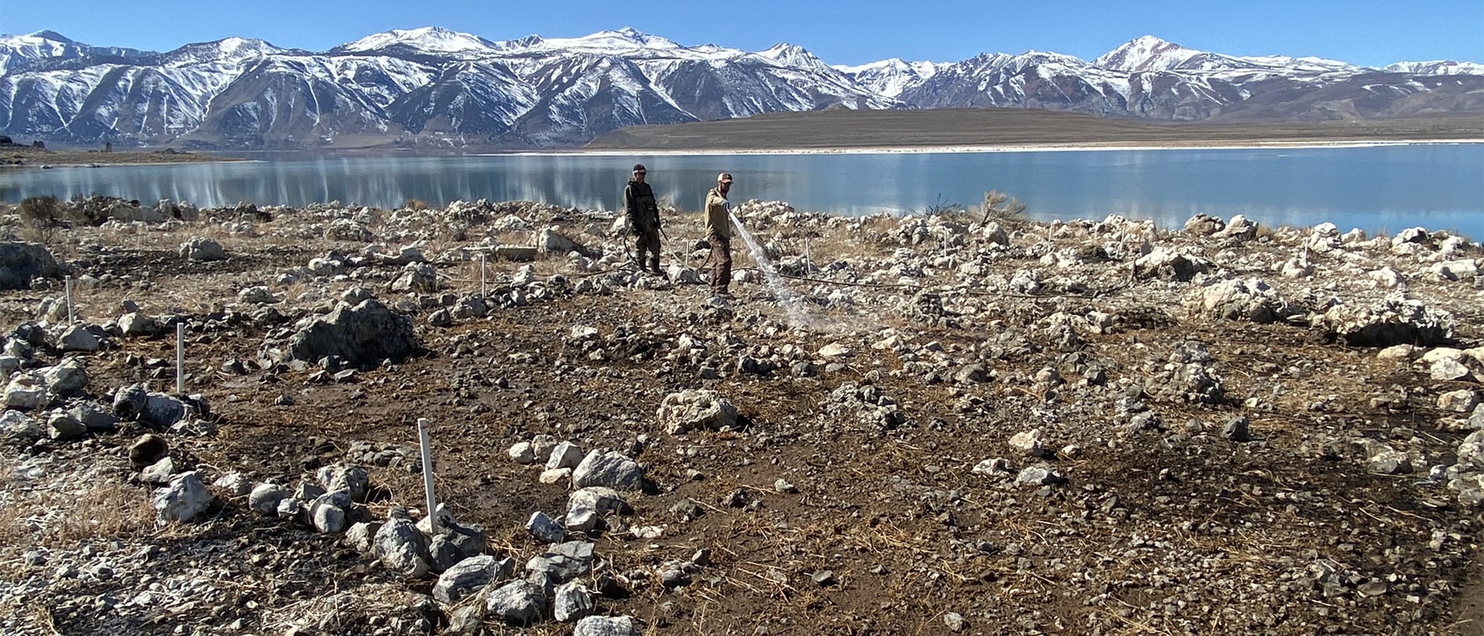 Two people stand in a bare rocky area on a lakeshore, one person is spraying water on the ground, a lake is in the background with snow capped mountains in the distance under a blue sky.