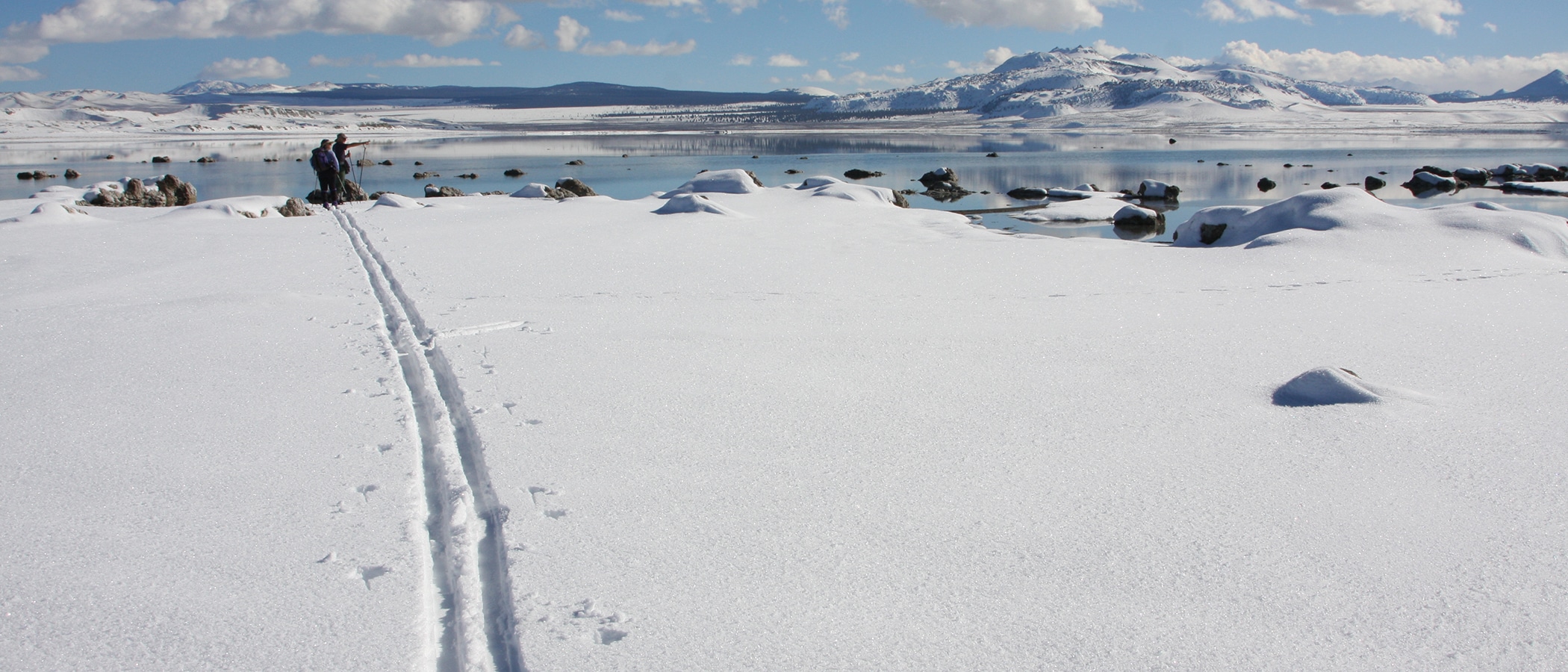 A single set of ski tracks cross fresh snow in the direction of tufa formations and a glassy lake surface reflecting snowy mountains and puffy white clouds.