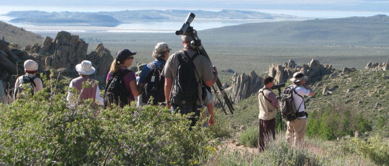 A group of eight people with scopes look in different directions into hilly sagebrush scattered with jutting gray rocks.