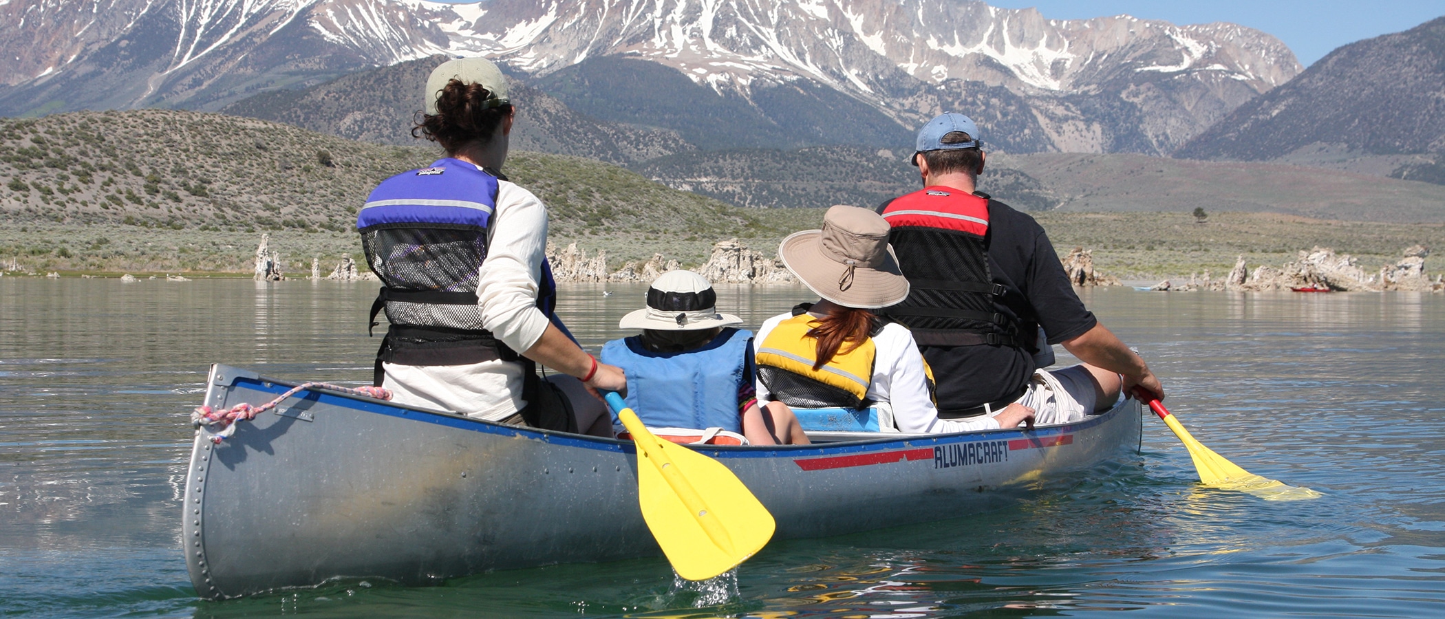 Four people paddle in an aluminum canoe with tufa formations and gray mountain peaks streaked with snow in the distance.