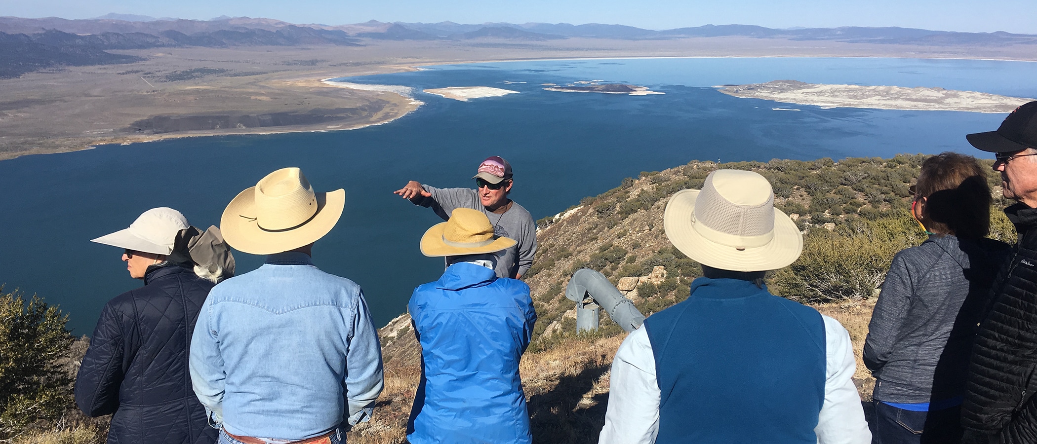 Six people gather around a guide who is using his hands to describe something, and the group is standing on a high precipice overlooking Mono Lake stretched out in a large expanse of blue with a clear view of the islands in the lake and the desert all around.