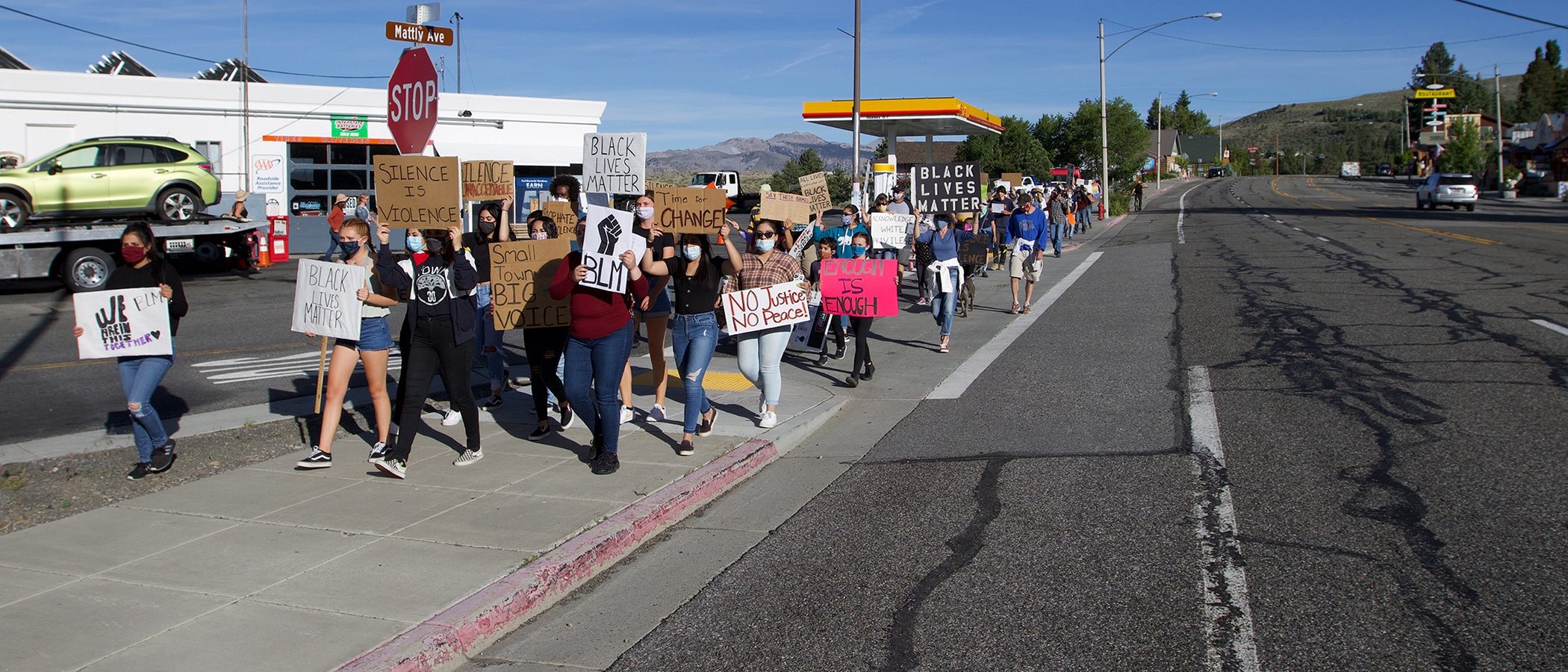 A large group of protestors walk down Mattly Ave with signs reading "Black Lives Matter" and "Enough is Enough". The Shell gas station is behind the group and there is a blue sky.