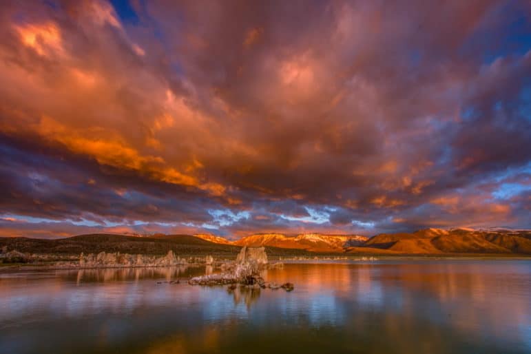 Stunning sunrise view at Mono Lake looking across the water, west to the Sierra Nevada which has golden light and dramatic stormclouds above a colorful reflection and tufa towers in the foreground.