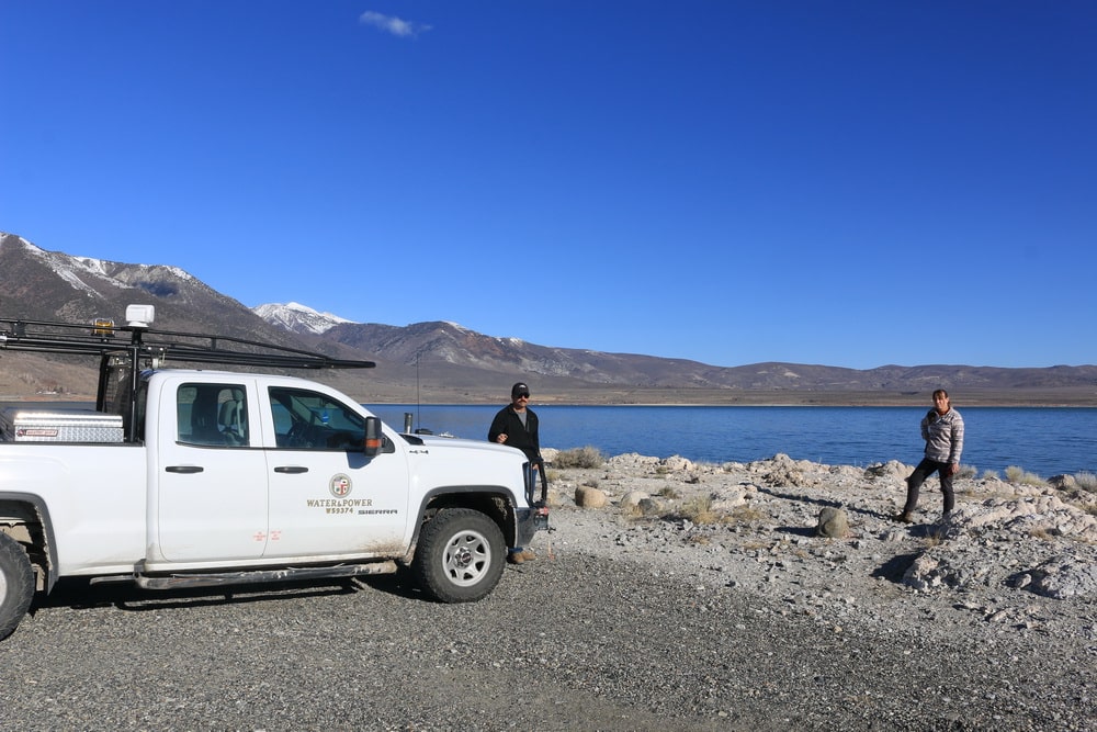 Two people stand on the rocky gray beach in front of a white truck which says "water and power". The sky is blue and cloudless and the water is bright blue.