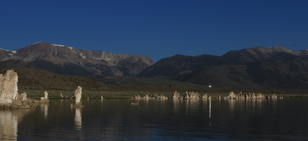 Glassy lake in front of Sierra Nevada with tufa towers reflecting in the morning light with one point of reflection in the lake from a mirror on a hill above.