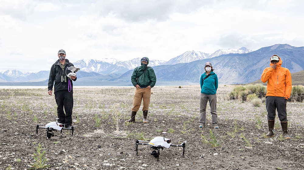 Four people wearing rain coats, boots, hats and masks, stand apart behind two drones which are sitting on a rocky ground. The snow capped Sierra Nevada mountains are behind them.