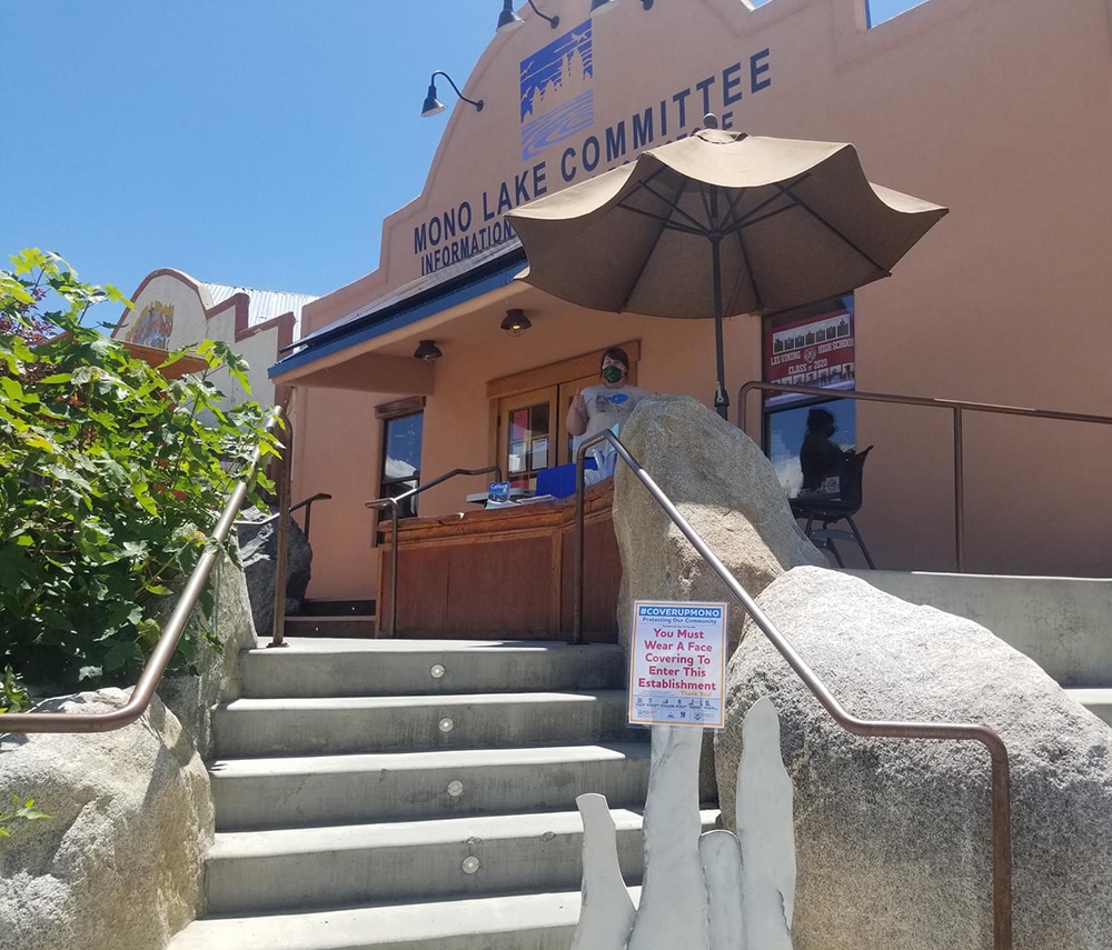 A woman in a face mask stands at a desk outside of the Mono Lake Committee building at the top of the stairs. There is an umbrella open to block the bright sun, and a small sign at the bottom of the stairs establishing Covid protocol. 