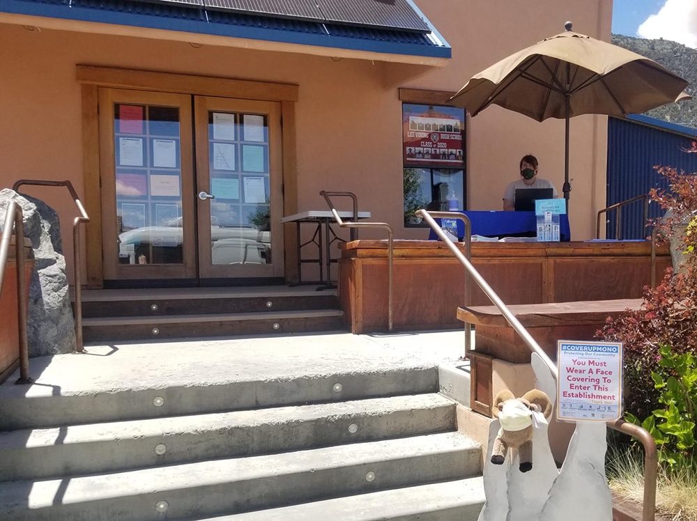 A woman wearing a mask sits outside the front of the Mono Lake Committee at a desk set up with papers. The doors to the store are closed and covered with paper signs. It is a sunny day.