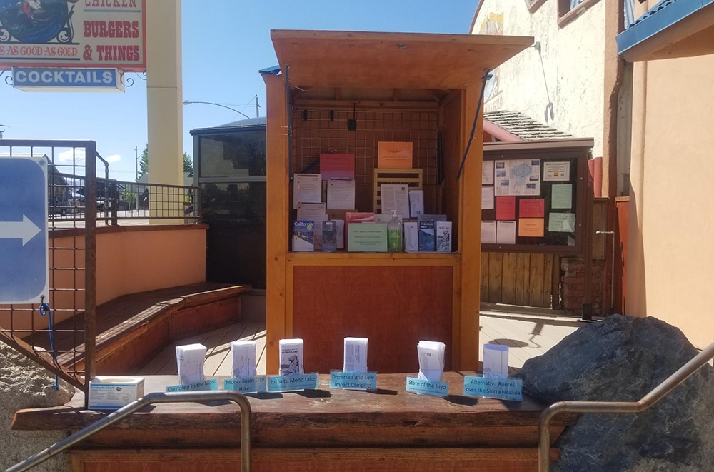 The left side of the information booth shows a wooden structure filled with colorful pamphlets and hand sanitizer. A table in front of the structure holds white paper maps and pamphlets labeled in blue.