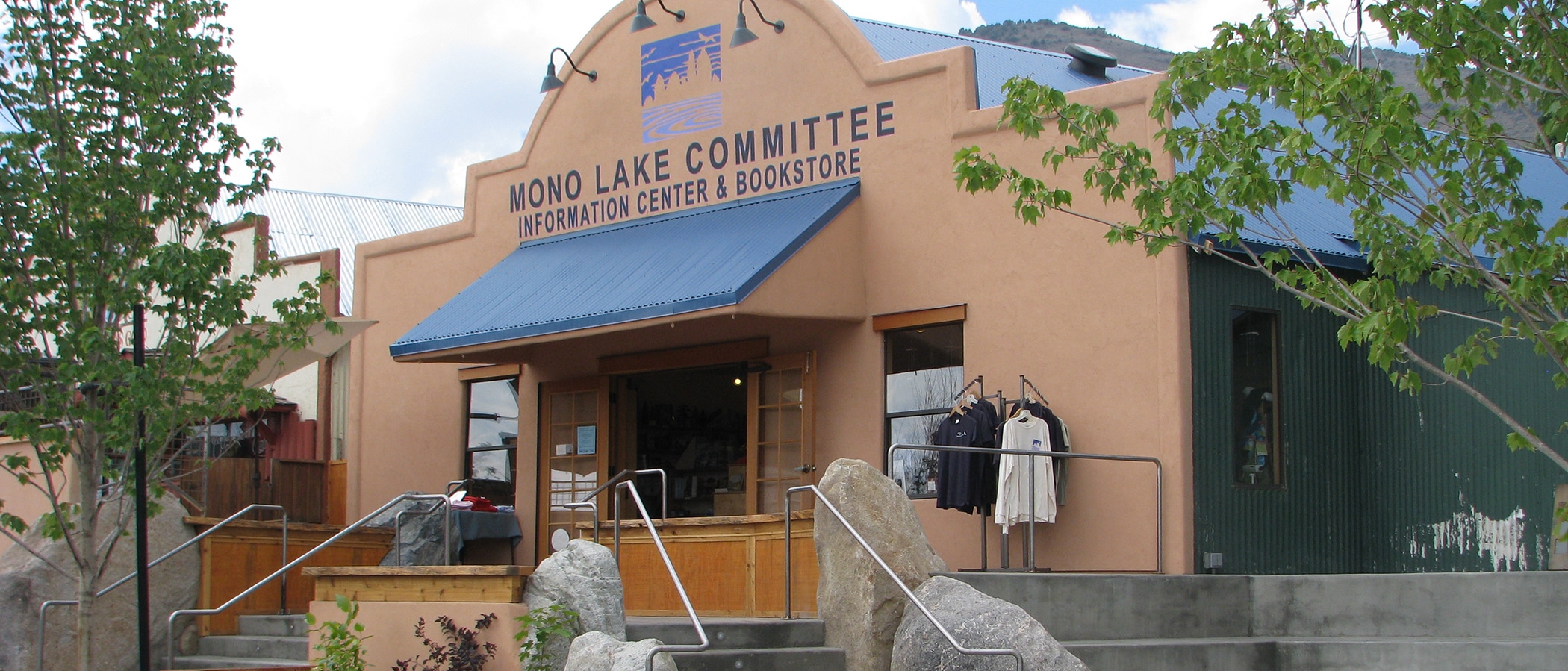 Mono Lake Committee Information Center & Bookstore storefront with a blue awning, trees, stairs, large landscaping rocks, and doors open to the public.