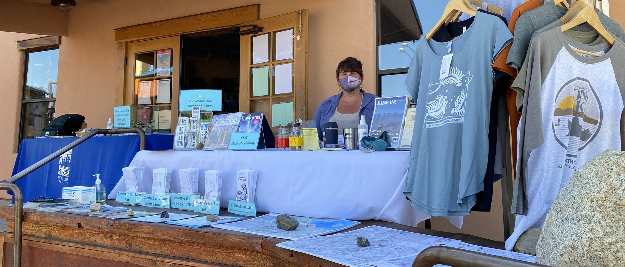 Woman in a face mask at a table set outside the Mono Lake Committee Information Center & Bookstore with information for visitors.