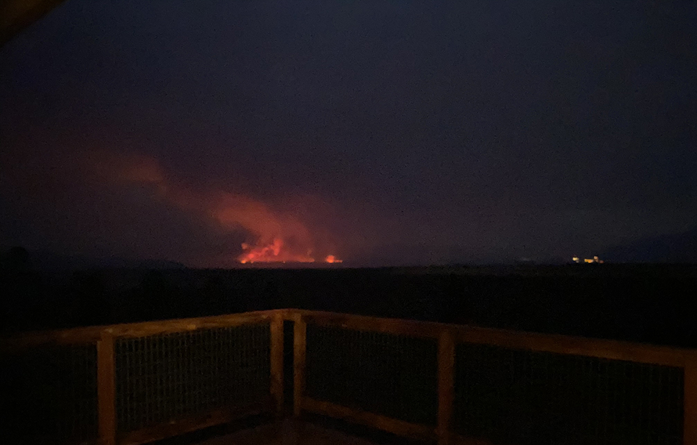 A pinkish glow with lighter pink smoke stand out against a dark sky and black land, with a wooden railing in the foreground.