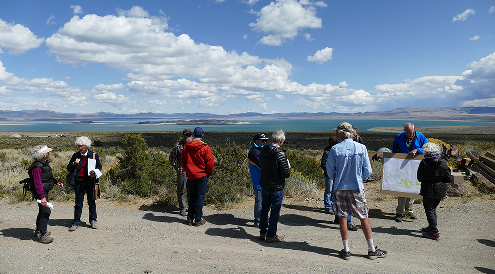 A group of several people survey the area on a gravel patch, looking out at the vast turquoise waters of Mono Lake. Some hold papers and point to maps. The sky is blue with clouds, and mountains stretch beyond the lake.