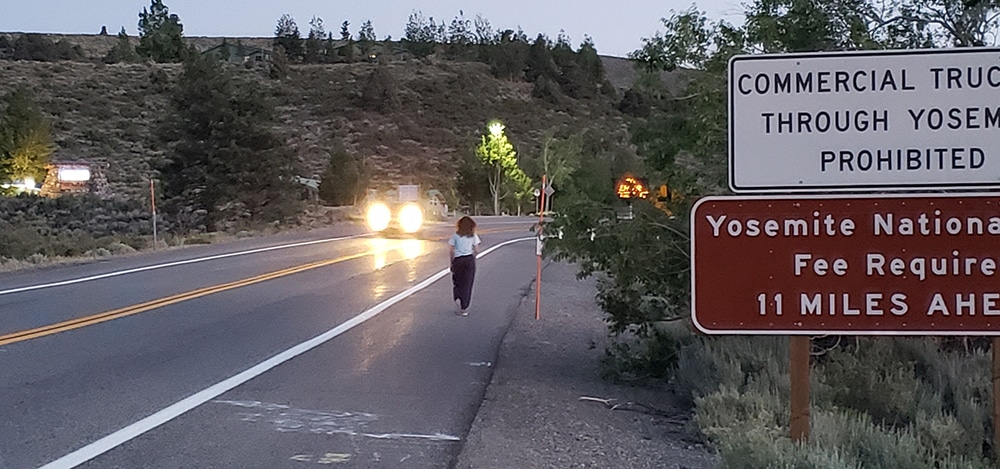 A pedestrian walks on the side of the highway at dusk, with a car's headlights flashing towards her.