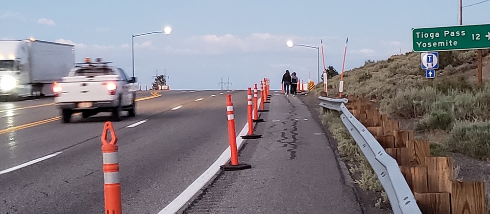 A white truck zooms past two pedestrians walking along the shoulder of the highway which is separated by orange cones.