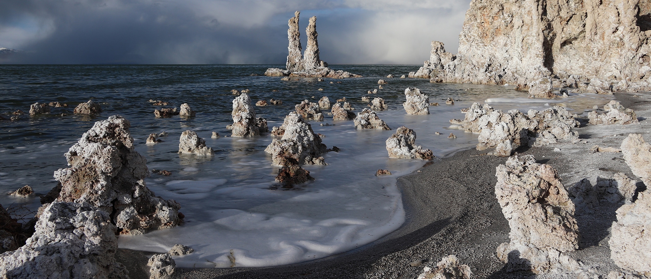 A storm brews in the distance behind a large grove of tufa towers on the shore of Mono Lake with a sandy peninsula dotted with smaller tufa mounds, in dramatic light, in the foreground.