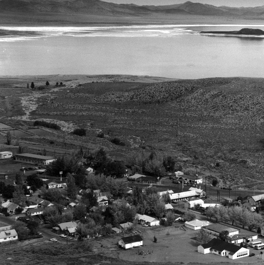 A black and white photo of the small town of Lee Vining, taken from a hill above, showing about 30 homes and shops with Mono Lake and black Negit Island and white Paoha Island, and the full moon rising in the distance.