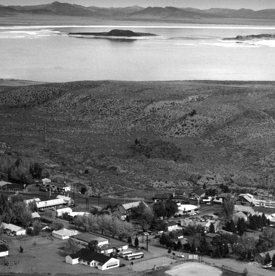 A black and white photo of the small town of Lee Vining, taken from a hill above, showing about 30 homes and shops with Mono Lake and black Negit Island and white Paoha Island, and the full moon rising in the distance.