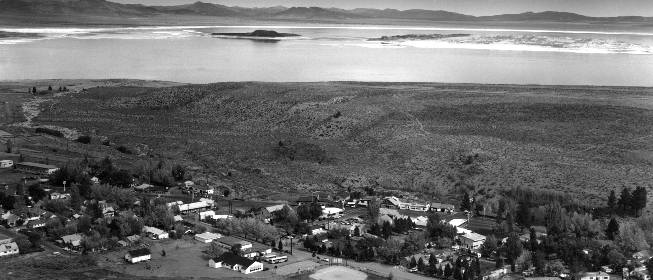 A black and white photo of the small town of Lee Vining, taken from a hill above, showing about 30 homes and shops with Mono Lake and black Negit Island and white Paoha Island, and the full moon rising in the distance.
