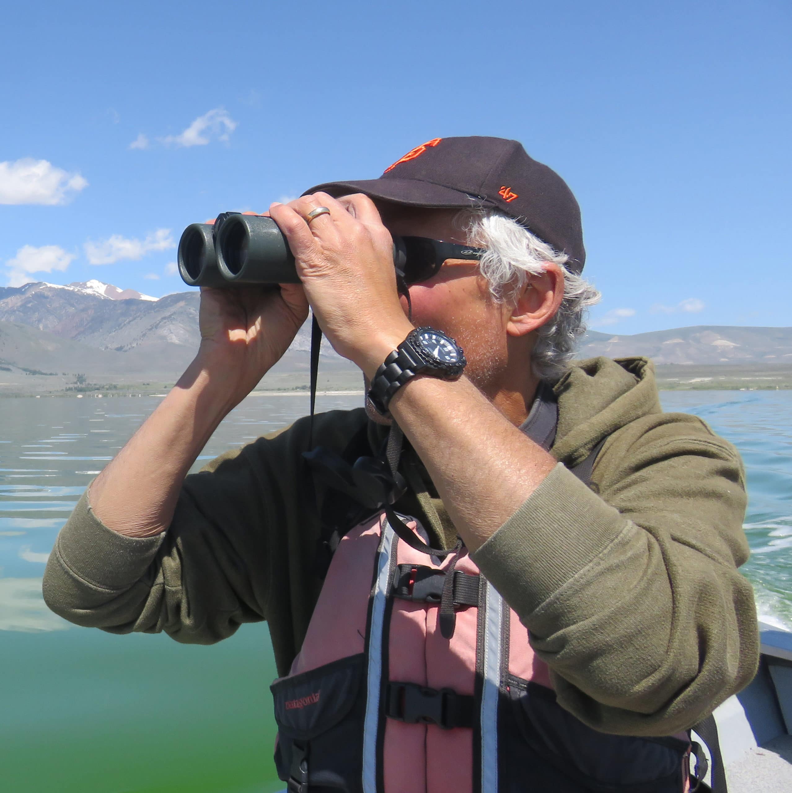 Researcher holds a pair of binoculars up to his eyes while wearing a life jacket and sitting on a boat with Mono Lake and mountains in the background.