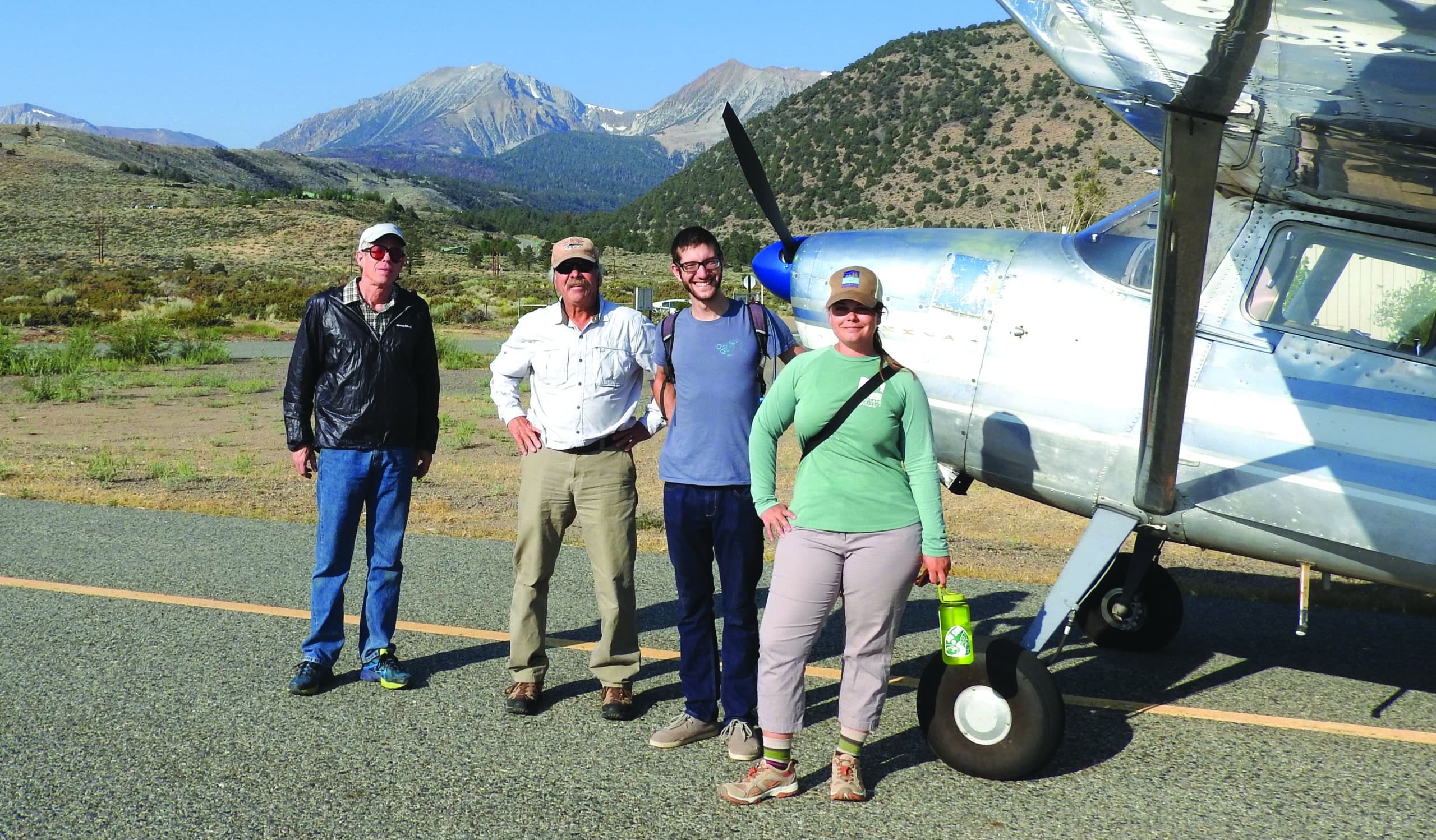 Four people stand in front of a single engine propeller plane on a small landing strip close to the mountains.