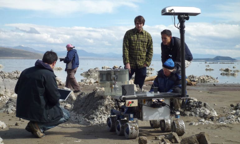 Four people on the shore of Mono Lake watching a prototype of the Mars Rover, a rolling platform with six wheels and various instrumentation, as it approaches a tufa mound.