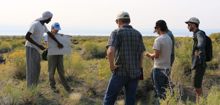 A group of six people standing outside in an area of open shrubs, with two people talking about and showing a chart while the others look on.