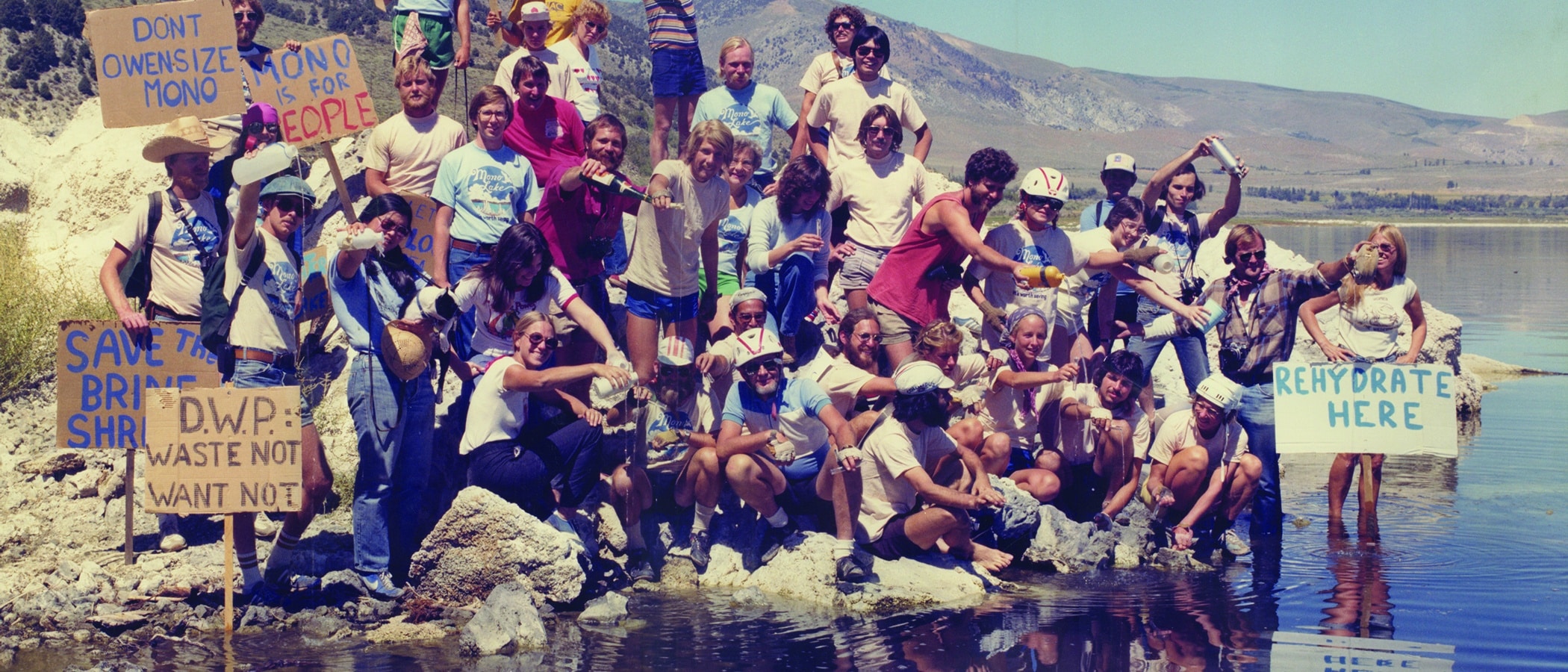 Group of about 50 people with hand-painted signs and Mono Lake T-shirts and many with bike helmets on pouring water from bottles and vials into Mono Lake.