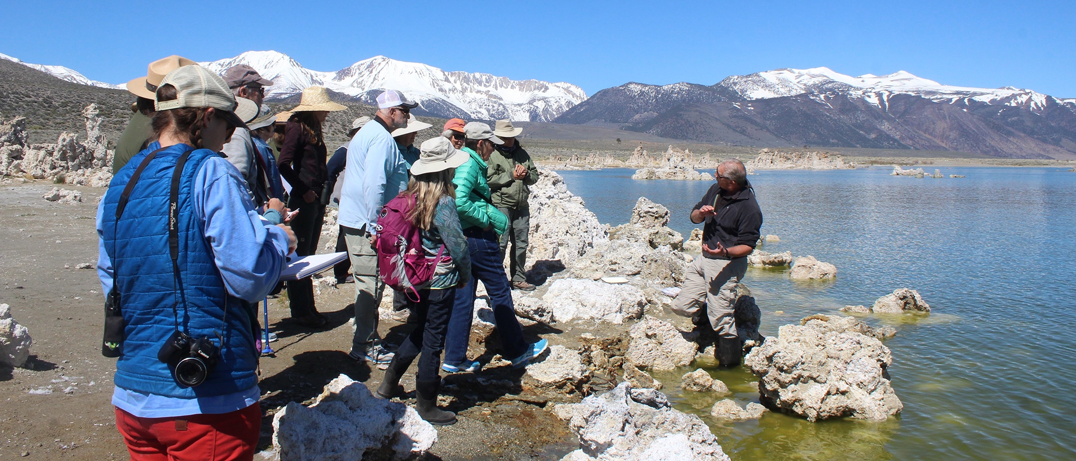 About 15 people standing on the Mono Lake shoreline listening to a person standing in Mono Lake leading a tour on a beautiful sunny day with the snowy Sierra Nevada in the background.