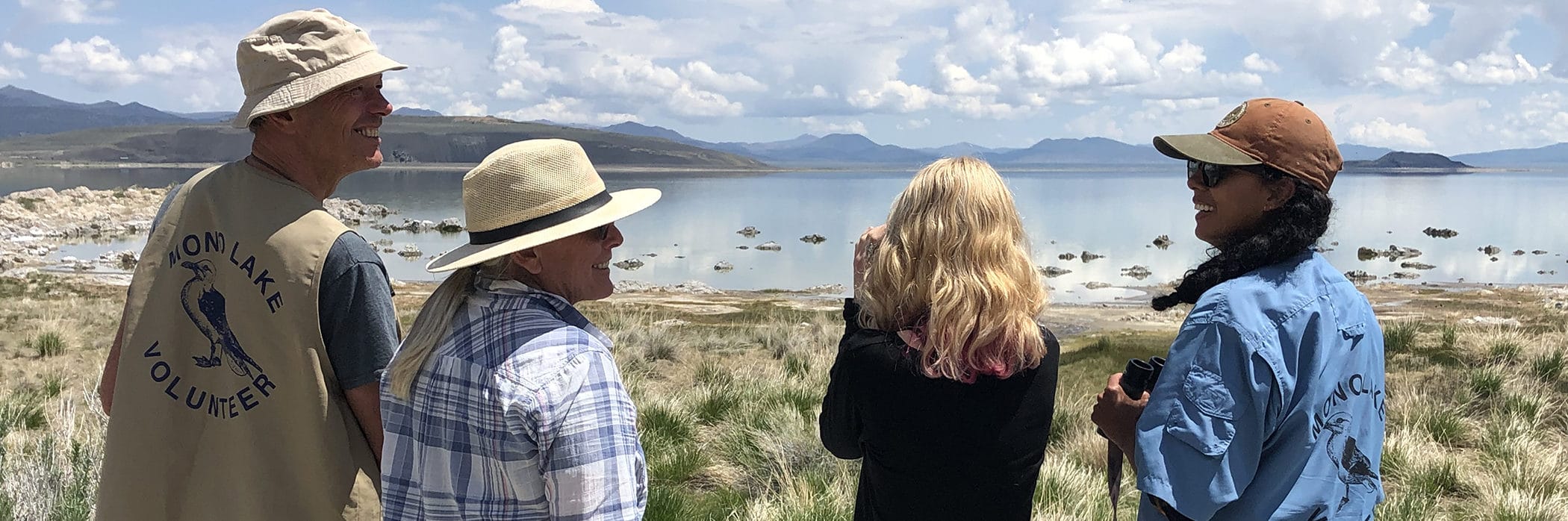 Four people overlooking Mono Lake on a bright sunny day smile at eachother while one looks through binoculars and tow of them have official Mono Lake Volunteer shirts and vests.