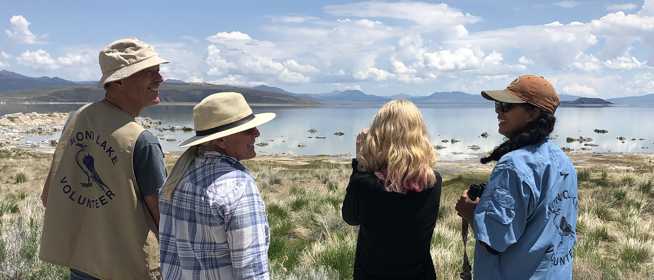 Four people overlooking Mono Lake on a bright sunny day smile at eachother while one looks through binoculars and tow of them have official Mono Lake Volunteer shirts and vests.