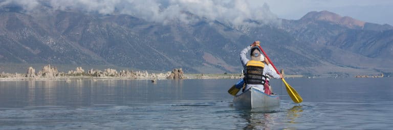 View from behind a person paddling a canoe on Mono Lake with tufa towers and tall mountains shrouded in clouds in the distance.